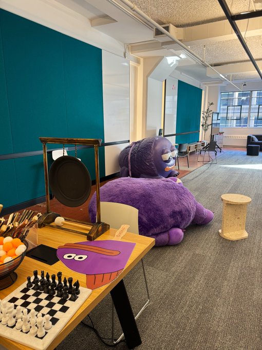 A giant purple mascot character rests on the floor of a colorful office lounge featuring a chessboard, gong, and playful desk items, capturing the whimsical "gmonad" aesthetic of the space.
