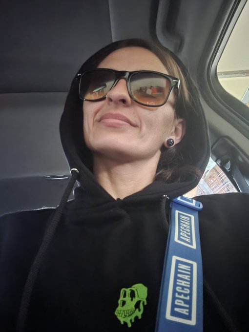 A selfie of a person wearing sunglasses and a black hoodie with a green dripping skull graphic, carrying an APECHAIN branded blue strap.