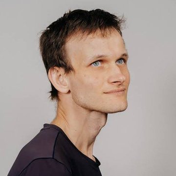 A portrait photo of a young man with short brown hair, blue eyes, and slight stubble, wearing a black t-shirt against a gray background, looking at the camera with a subtle smile.