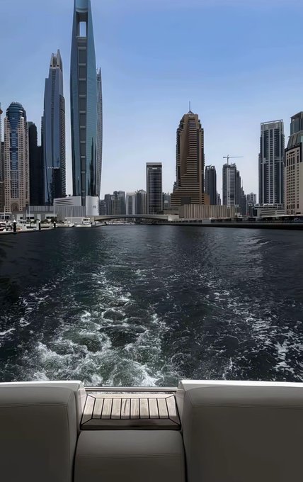 A boat glides through the Dubai Marina, leaving a wake behind as it passes by the towering skyline including the iconic Cayan Tower. The image pairs with a motivational "GM CT" message encouraging focus and success in trading.