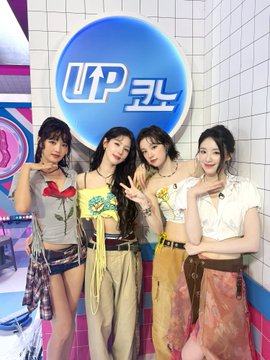 Four women posing in front of a circular blue and white 'UP' logo on a white tiled wall, wearing varied colorful outfits including a tulip top, a yellow top, a white ruffled top, and an orange skirt.