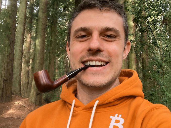 The image features a man wearing an orange hoodie with a pipe sticking out of his mouth. The man has a smile on his face as he poses for the photo, likely sharing a moment or expressing gratitude towards someone named Casey Rodarmor. The background includes trees and bushes, suggesting that they are in a forest setting.