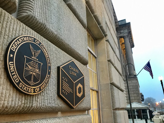 A photograph of two plaques mounted on a stone building facade: a circular seal for the 'Department of Commerce United States of America' and a hexagonal 'Chain Link' symbol plaque, with an American flag visible in the background.