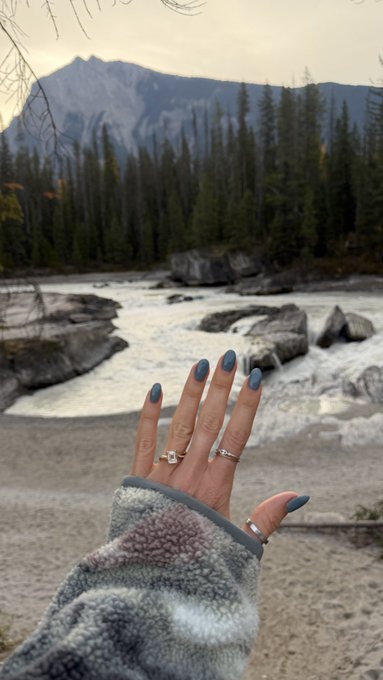 A hand with blue-painted nails and silver rings is extended toward a rushing river with rocky banks, evergreen trees, and a mountain in the background.