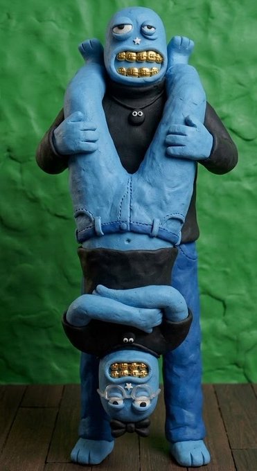 A surreal blue clay sculpture depicting a stacked figure with gold teeth - the top figure wears a black shirt with a small face design, and the bottom inverted figure wears glasses and also displays gold teeth, set against a green backdrop.