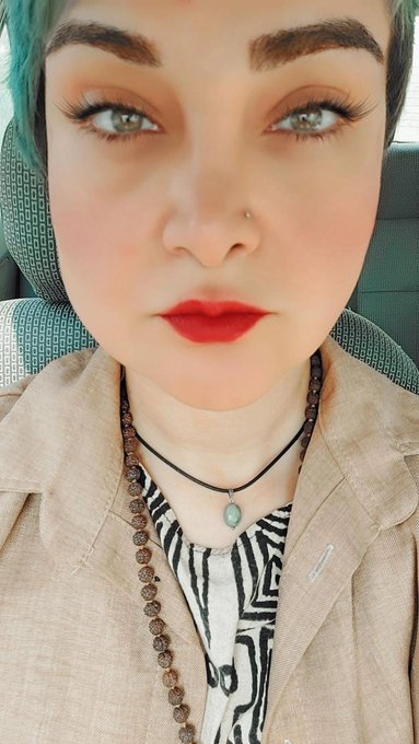 Close-up selfie of a person with bold makeup including red lipstick, heavy eyeshadow, defined brows, and a nose ring, wearing a tan blazer over a patterned shirt with a beaded necklace and pendant.