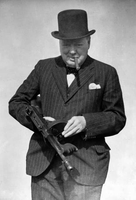 A black and white vintage photograph of a smiling man, resembling Winston Churchill, wearing a pinstripe suit, top hat, and bow tie, holding a submachine gun and a cigar.