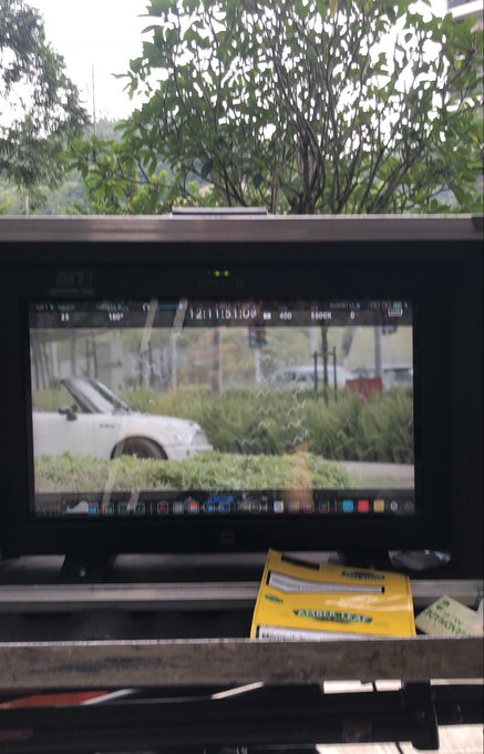 An outdoor camera monitor displays a live feed of a white car with its hood up parked near some bushes, with trees and overcast sky visible in the background behind the monitor.