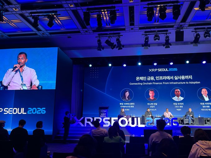 A panel discussion at 'XRP SEOUL 2026' titled 'Connecting Onchain Finance: From Infrastructure to Adoption' with four Korean-speaking panelists, while a man speaks into a microphone displayed on the large screen to the left.