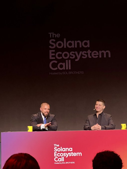 Two men in suits seated at a panel for 'The Solana Ecosystem Call,' hosted by SOLBROTHERS, with the event name displayed on the backdrop and podium.
