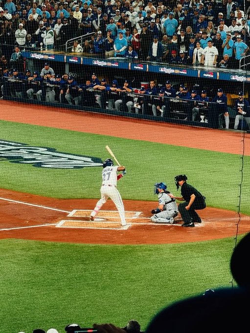 A baseball game in progress shows a batter in a white uniform (#77) at home plate with the catcher and umpire behind him, while spectators in teal shirts fill the stands above MLB.com signage.