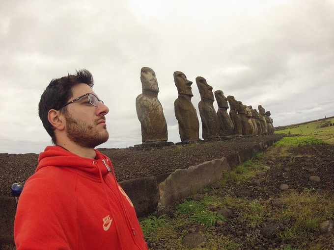 A young man in a red Nike hoodie and glasses standing in profile before a row of Moai statues at Easter Island under an overcast sky.