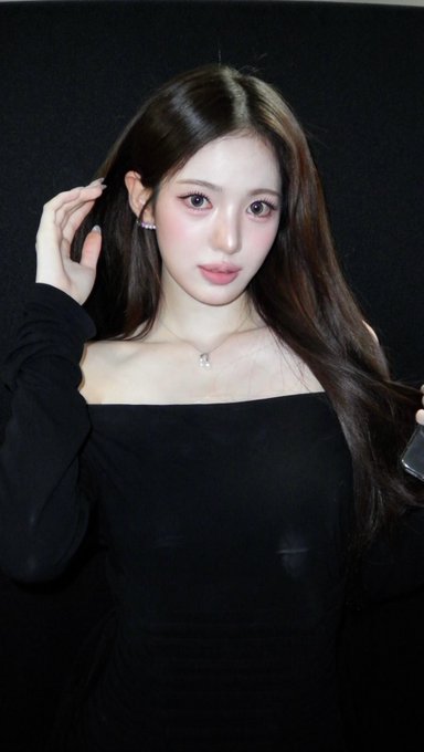A young woman with long brown hair poses for a selfie, wearing a black off-shoulder top and a delicate necklace.