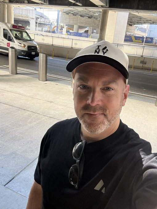 A man wearing a black NY cap and a black Adidas t-shirt takes a selfie at what appears to be an airport or transportation hub.