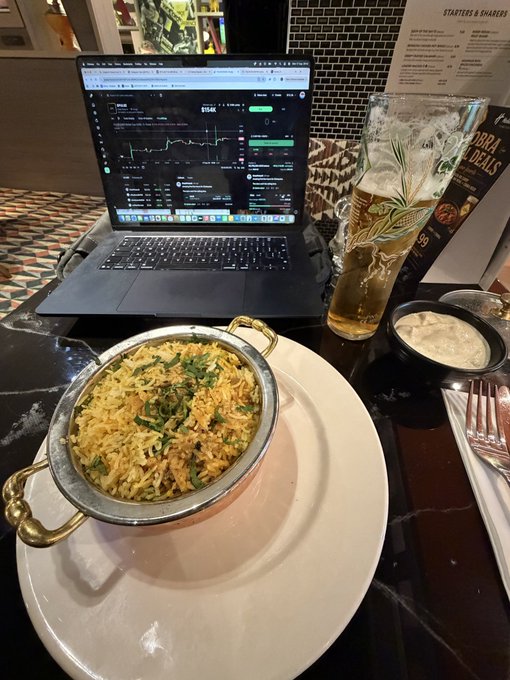 A meal at a restaurant featuring a dish of biryani in a copper pot, a glass of beer, and a small bowl of sauce on the table, with a laptop showing a stock or crypto trading chart in the background.