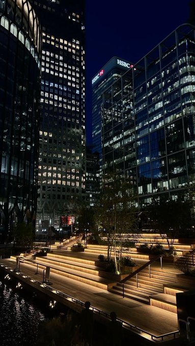 A nighttime cityscape shows illuminated modern skyscrapers, including a building with an 'HSBC' sign, towering over a lit outdoor amphitheater.