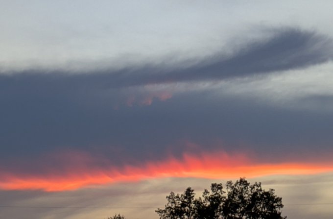 A dramatic sunset photograph showing dark storm clouds with a vibrant red and orange glow illuminating the sky above silhouetted tree tops.