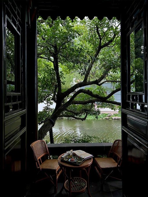 A serene view through an open doorway of a traditional Chinese pavilion, looking out onto a lush green tree and pond, with a small table set with tea between two rattan chairs.