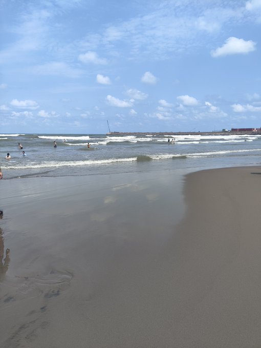 A serene beach scene with people wading in the shallow waves under a blue sky dotted with white clouds, with footprints visible on the sandy shore in the foreground.