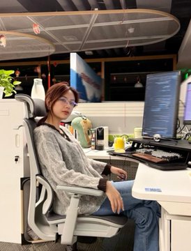 A woman sits in an ergonomic office chair beside a computer setup with dual monitors, apparently resting in a home workspace, which ties to the post about a 24-year-old who transformed her second bedroom into a home server room. The monitors display lines of code or text, suggesting she's working on or managing her Mac mini-based cryptocurrency mining or computing operation that earned her $58,400.