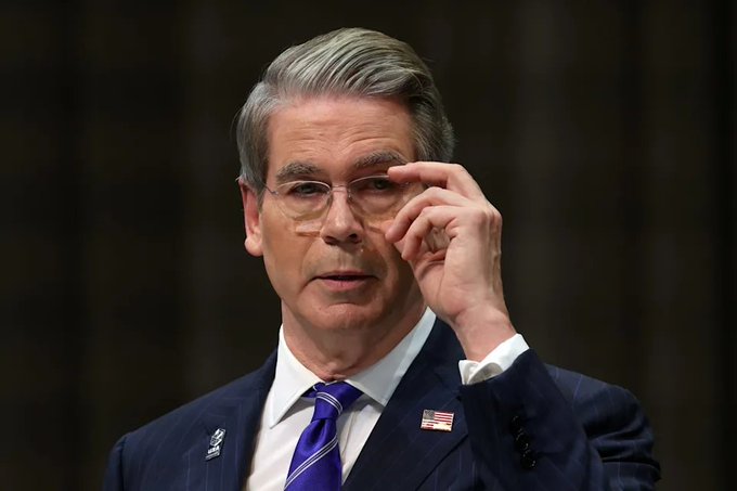 A photograph of a gray-haired man in a dark pinstripe suit, white shirt, and purple tie adjusting his glasses, with American flag and US flag pins on his lapels.