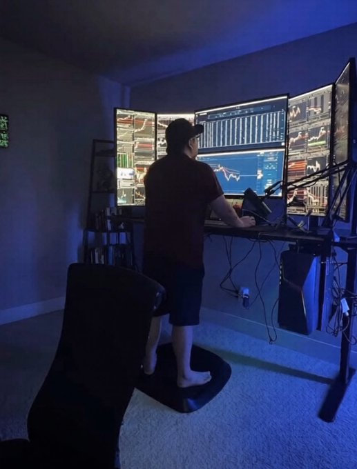 A person stands at a multi-monitor trading setup in a dimly lit room with blue lighting, analyzing various financial charts and data displayed across three screens.