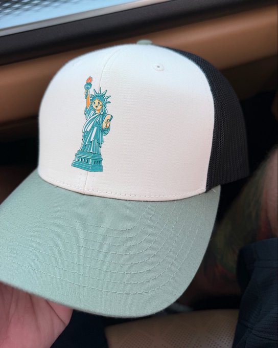 A white and light green trucker hat with a black mesh back features an embroidered Statue of Liberty design, held in someone's hand inside a car.