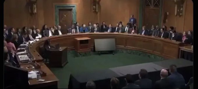 A wide shot of a government hearing room with officials seated around a curved wooden desk with a witness table and TV screen in the center.