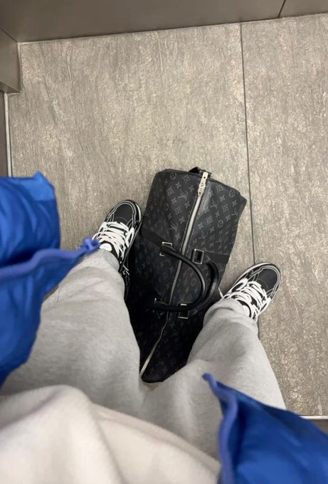 A first-person view looking down at someone's legs in gray sweatpants and black and white sneakers, standing on a tiled floor next to a Louis Vuitton monogram duffel bag and a blue jacket.