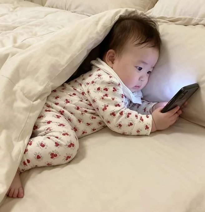 A baby in a floral onesie lying on a bed under covers, intently looking at a smartphone screen.