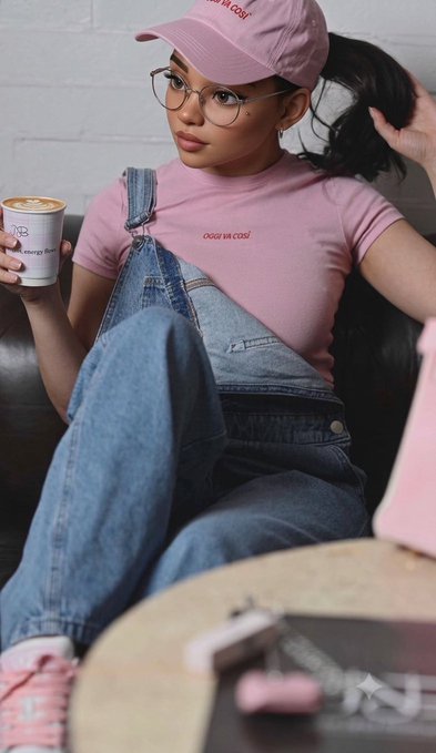 The image features a woman sitting on a couch, wearing a pink shirt with a cap and glasses. She has her hair pulled back into a ponytail. In front of her are two cups - one white and one red. The background shows a brick wall and a table. There is also a keychain visible in the scene.