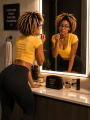 A woman with dreadlocks wearing a yellow top and black pants stands facing a mirror in a bathroom, applying makeup.