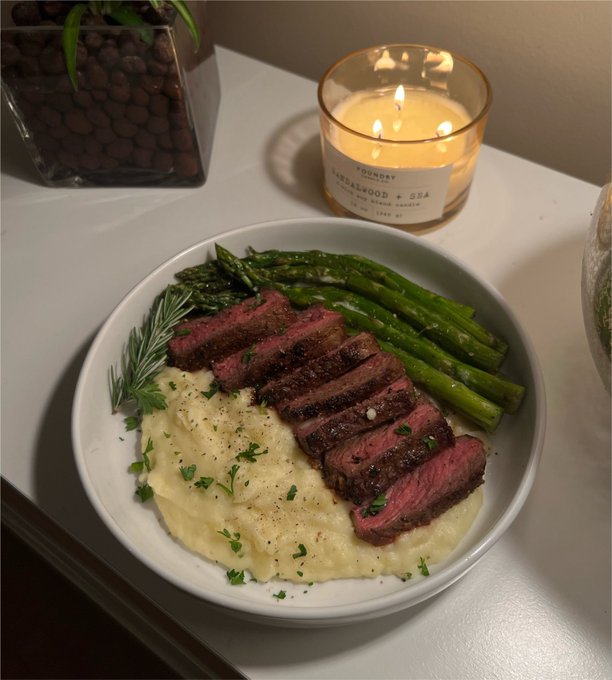 A bowl of sliced medium-rare steak served over creamy mashed potatoes with asparagus and fresh herbs, set next to a lit candle labeled 'FOUNDRY SANDLEWOOD + SEA' and a glass vase with clay pebbles.