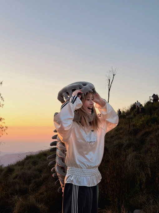 Young woman wearing a white jacket and track pants posing with a large caterpillar or centipede-like sculpture on her head against a sunset sky.