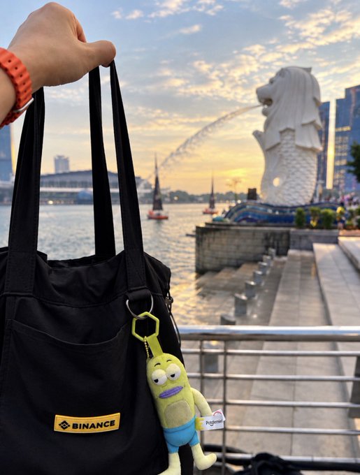 A hand holds a black Binance tote bag with a plush toy keychain attached, photographed against the Merlion statue at Marina Bay, Singapore.