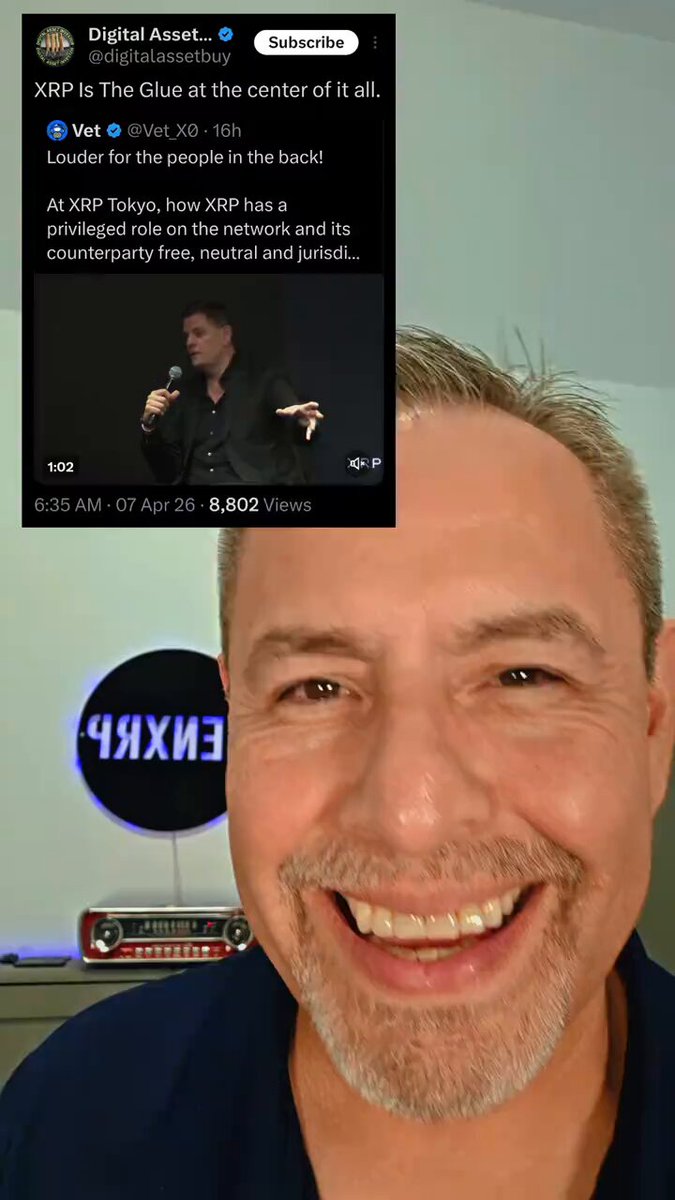 The image features a man with a beard, wearing a blue shirt, smiling at the camera while holding a microphone. The background of the photo shows a white wall with a sign that reads "XRP is the Glue at the center if it all!". This suggests that the man might be discussing or promoting XRP as an essential cryptocurrency for various purposes such as securing digital assets and transactions.