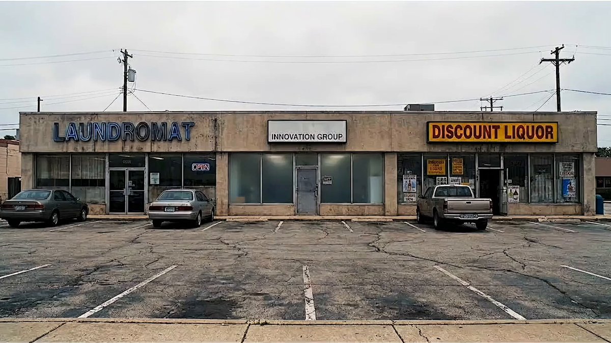 The image shows a large, empty parking lot with several cars parked in front of a building that has a sign for "Laundromat". The building appears to be an old business or store, possibly a laundromat, given its name. There are also two other buildings visible in the background, one on each side of the main building.