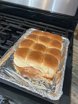 A tray of freshly baked sliders with golden-brown rolls sitting on aluminum foil on top of a stove burner, with visible meat filling between the buns.