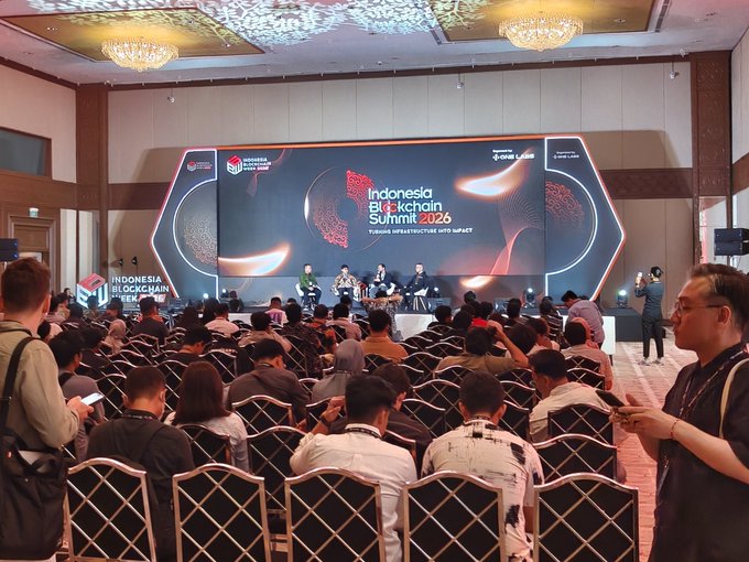 A photo of an audience seated in a large conference hall watching a panel discussion at the 'Indonesia Blockchain Summit 2026', with branded screens visible on stage and 'INDONESIA BLOCKCHAIN WEEK' signage to the left.