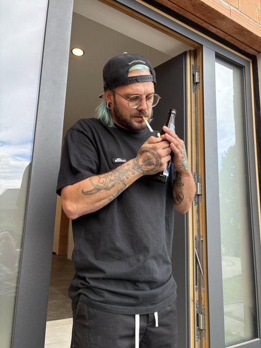 A man with tattooed arms, wearing a black cap, glasses, and a black Adidas t-shirt, stands in a doorway lighting a cigarette while holding a bottle.
