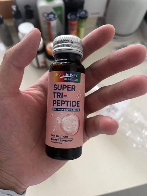 A hand holds a small dark glass bottle labeled 'Holistic Way Bio-Elixir SUPER TRI-PEPTIDE Collagen Shot 15,000MG Skin Solutions Dietary Supplement'.