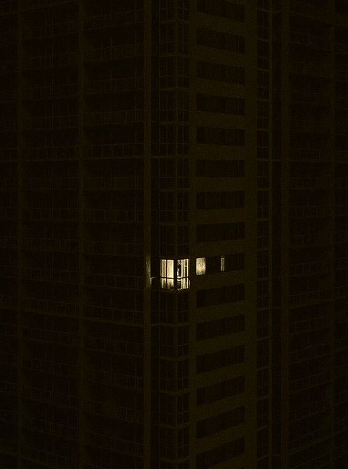 A single window shines faintly amid the vast dark facade of a high-rise building at night, embodying "the last light standing" as the only illuminated unit among countless darkened apartments. The solitary glow evokes a quiet, solitary presence within an otherwise pitch-dark tower.