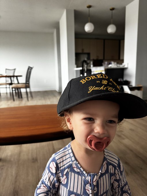 A young child wearing a black cap with 'BORED Yacht Club' in yellow text and a pacifier smiles in a modern apartment with a kitchen and dining area in the background.