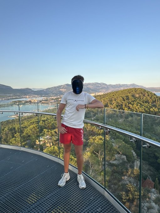A person wearing a white t-shirt, red shorts, and white sneakers stands on a glass-railed observation deck with a scenic view of mountains, coastline, and forest below.