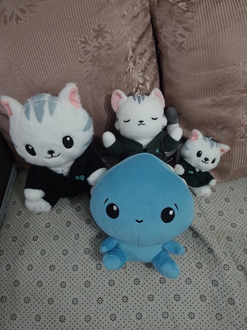 A group of four plush toys sitting on a couch: three white cats with blue stripes wearing black outfits and a round blue octopus character in the center.