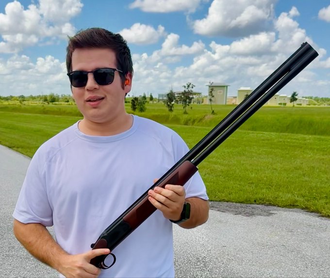 A man wearing sunglasses and a light purple t-shirt stands outdoors holding a wood-finished shotgun against a backdrop of green fields and a partly cloudy sky.