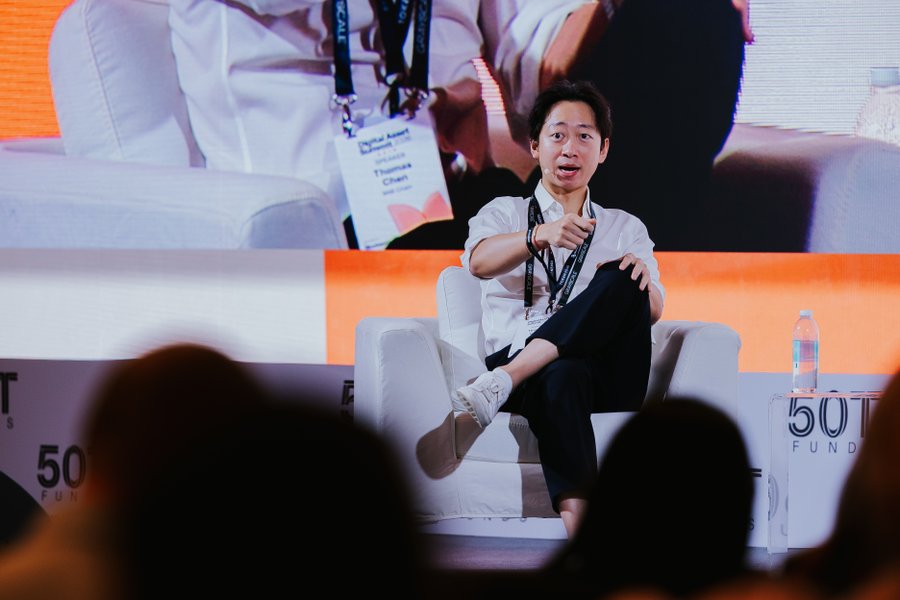 A man in a white shirt and dark pants sits on a light gray armchair on stage, gesturing while speaking at a panel discussion. Behind him, a large screen displays his image with a "50 Fund" logo and a name badge reading "Thomas Chen," matching his participation in a tokenization discussion at Blockworks DAS alongside representatives from Grayscale and S&P Global Ratings.