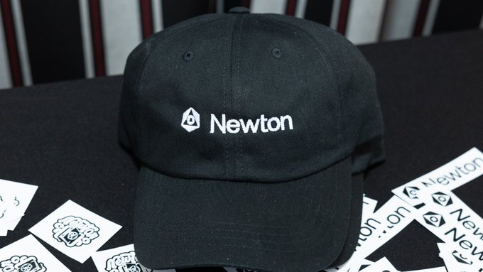 A black baseball cap with the white logo and text 'Newton' embroidered on it, sitting on a dark surface surrounded by white stickers bearing the same branding.