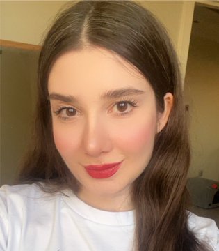 A close-up selfie of a young woman with long brown hair, prominent blush, and red lipstick, wearing a white top against a plain background.
