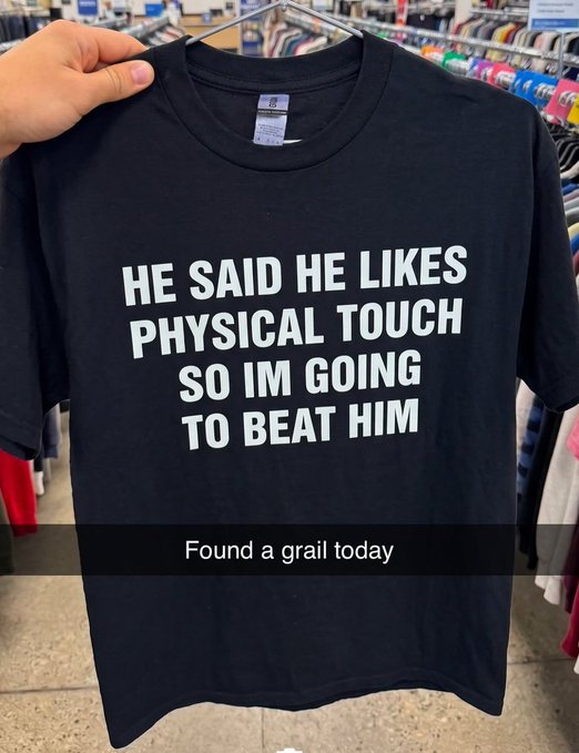 A black t-shirt held up in a store displays white text reading 'HE SAID HE LIKES PHYSICAL TOUCH SO IM GOING TO BEAT HIM,' with a caption overlay that says 'Found a grail today.'